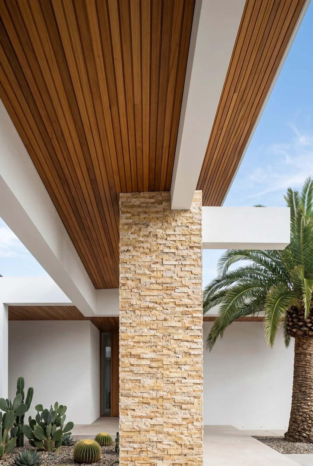 Exterior column faced in Scabos travertine ledger panel under a white soffit and timber ceiling, with palms against the sky behind.