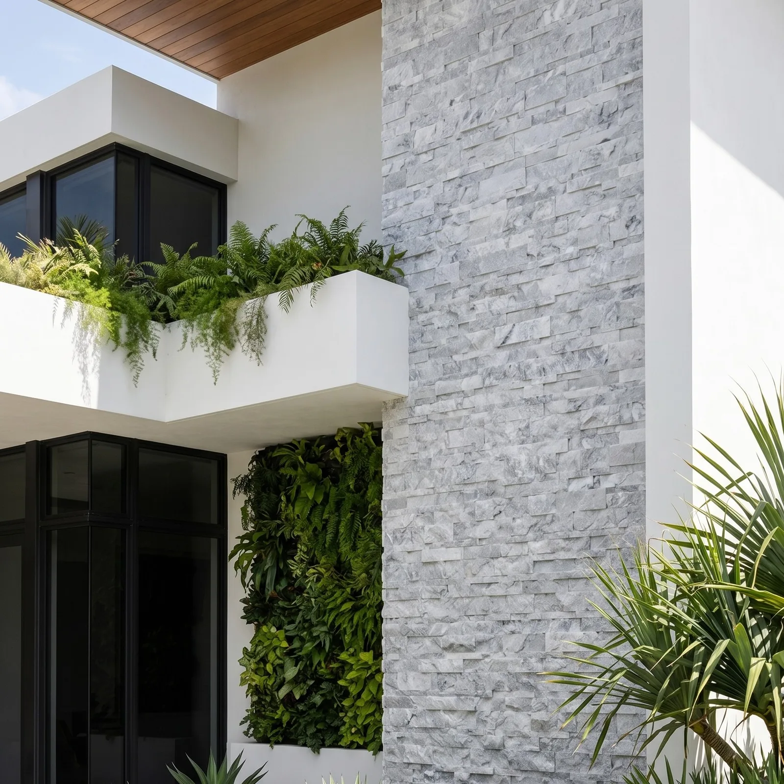 A house corner faced in Tundra Grey marble ledger panel, dark glazing to one side and a planted balcony spilling greenery over the pale grey stone.