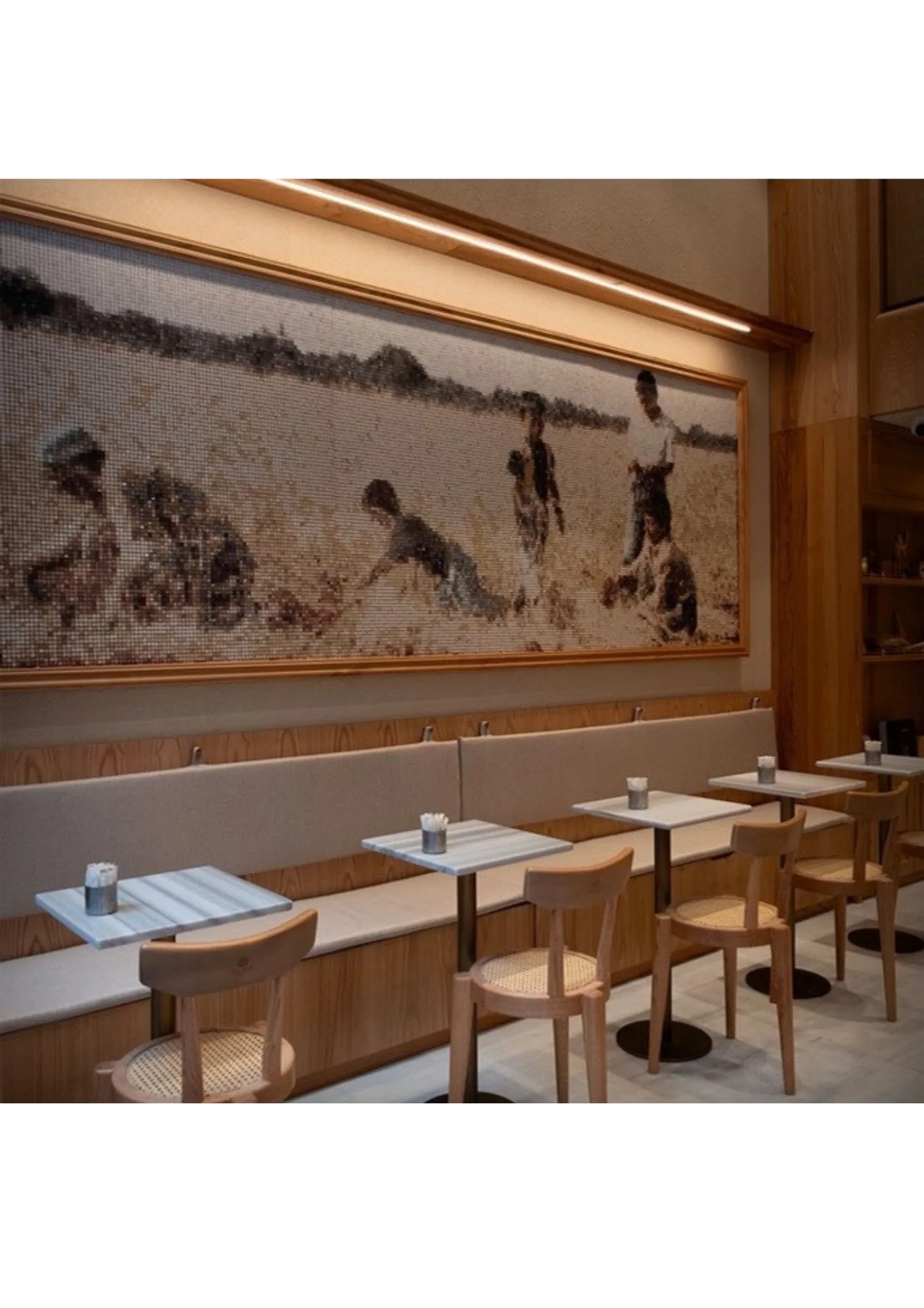 A Mosamia mosaic mural running the length of a café wall above banquette seating in a light timber frame, lit by a warm linear fixture, with marble-topped tables and cane chairs below.