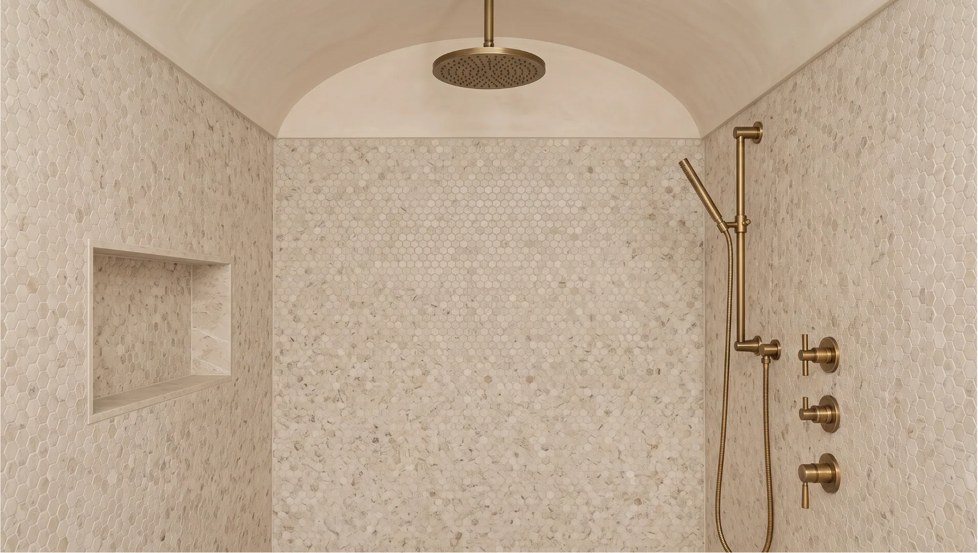 A shower alcove lined floor to vault in pale pebble mosaic, with a recessed niche and brushed brass fittings.