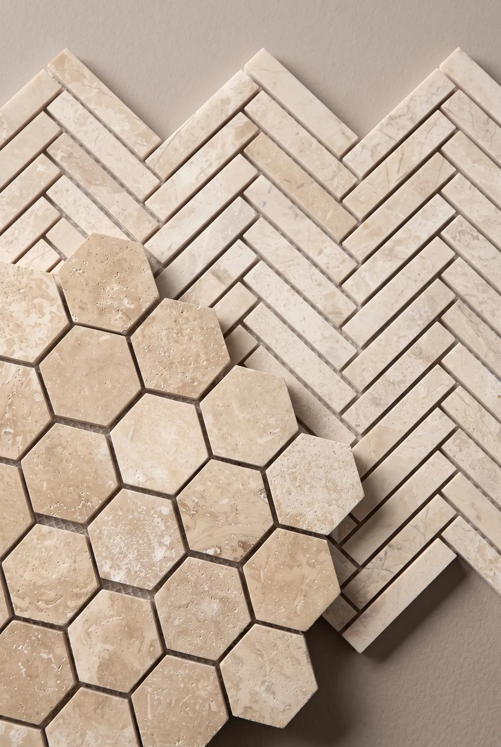 Two mosaic sheets overlapping on a neutral ground: a travertine hexagon mosaic laid across a cream marble herringbone, both in warm stone tones.