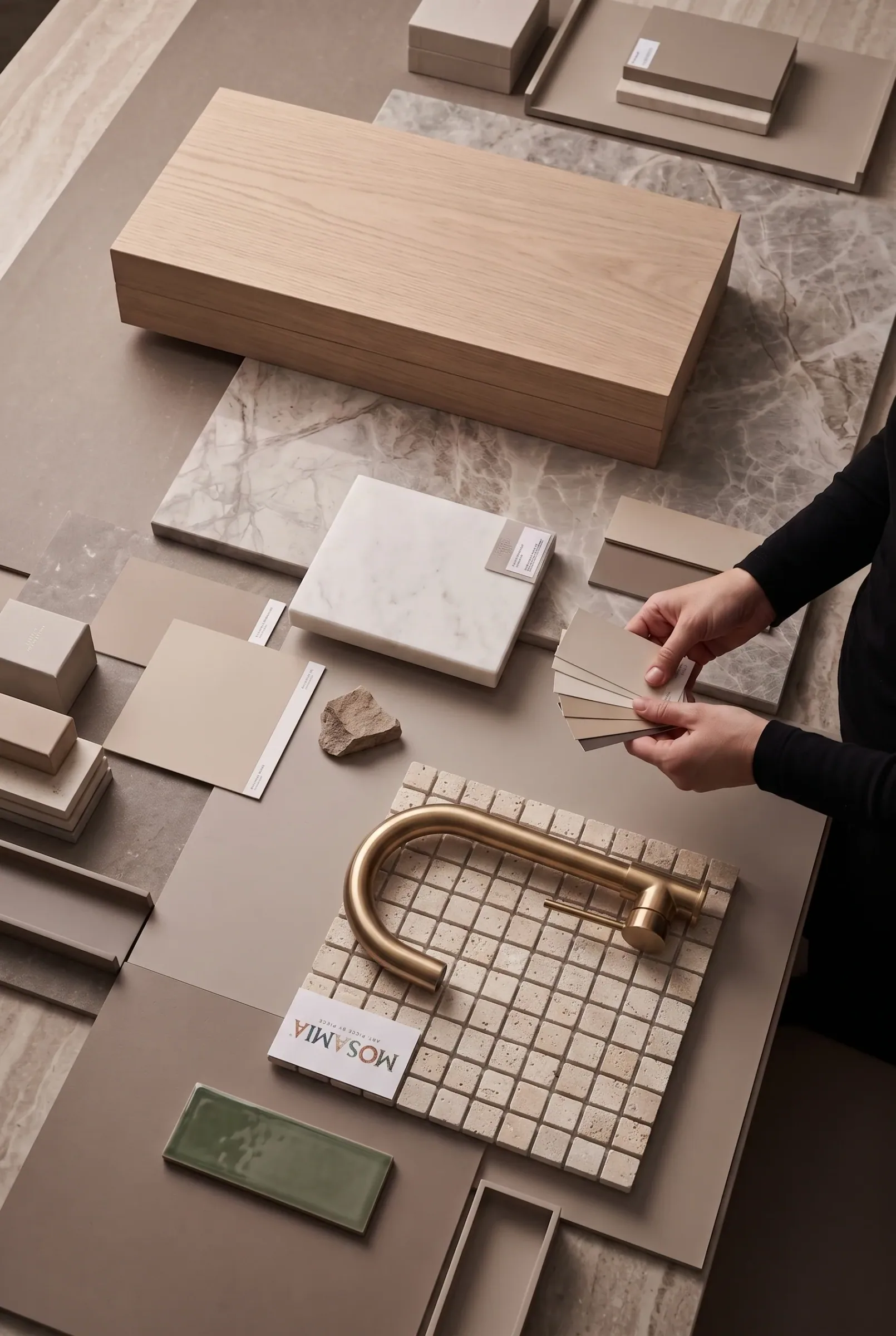 A specification desk seen from above: a timber sample box, marble and travertine tiles, paint chips fanned in a hand, a travertine mosaic sheet with a brass tap laid across it, and a Mosamia card.