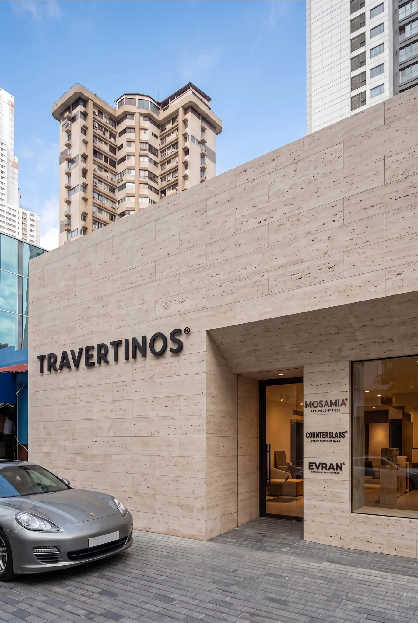 The same building from the street, its travertine elevation carrying the Travertinos name in raised lettering, with the Mosamia, Counterslabs and Evran marks on the entrance glass and residential towers rising behind.