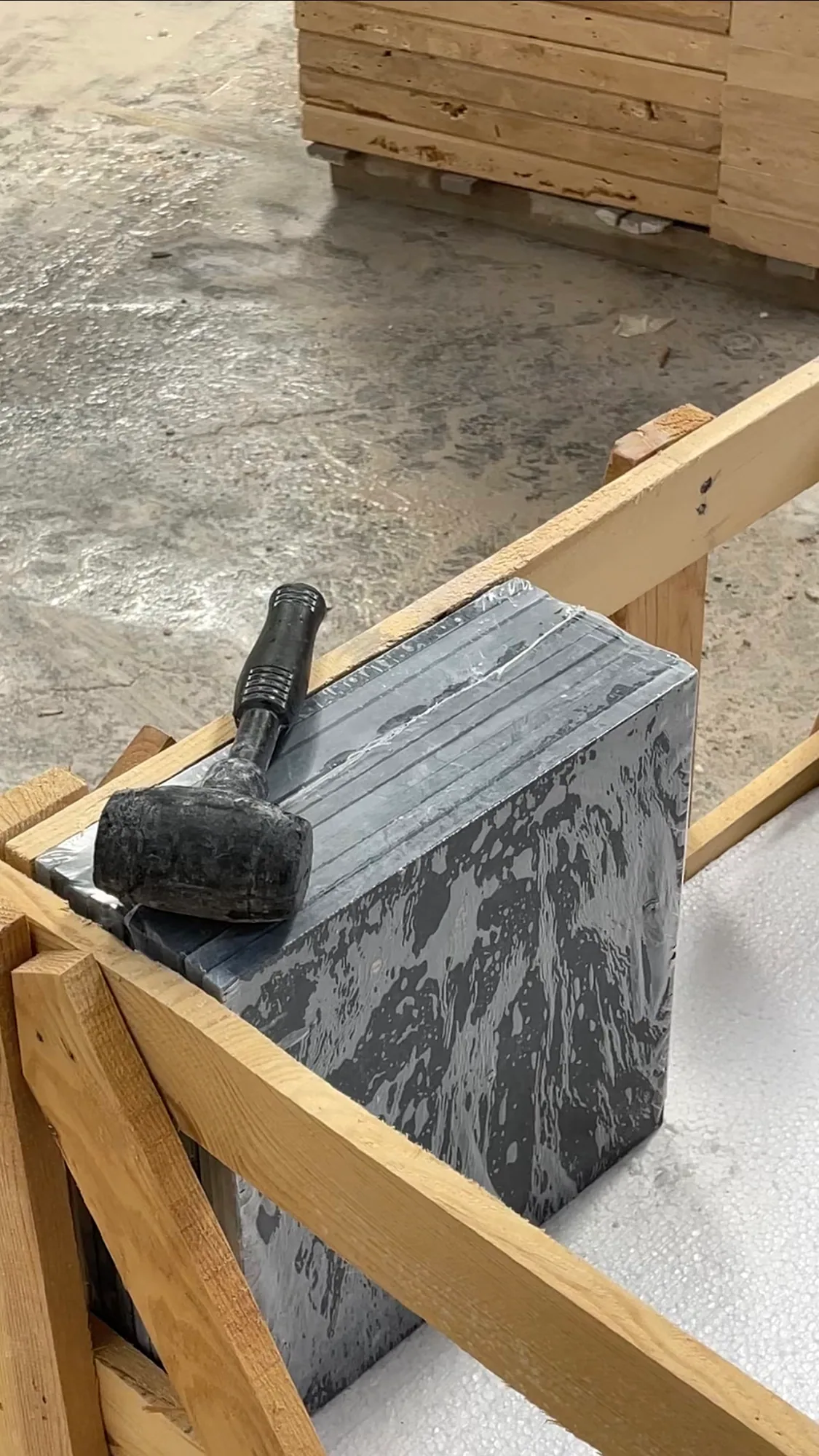 A rubber mallet resting on a shrink-wrapped bundle of dark grey stone tiles seated in a timber crate on the warehouse floor.
