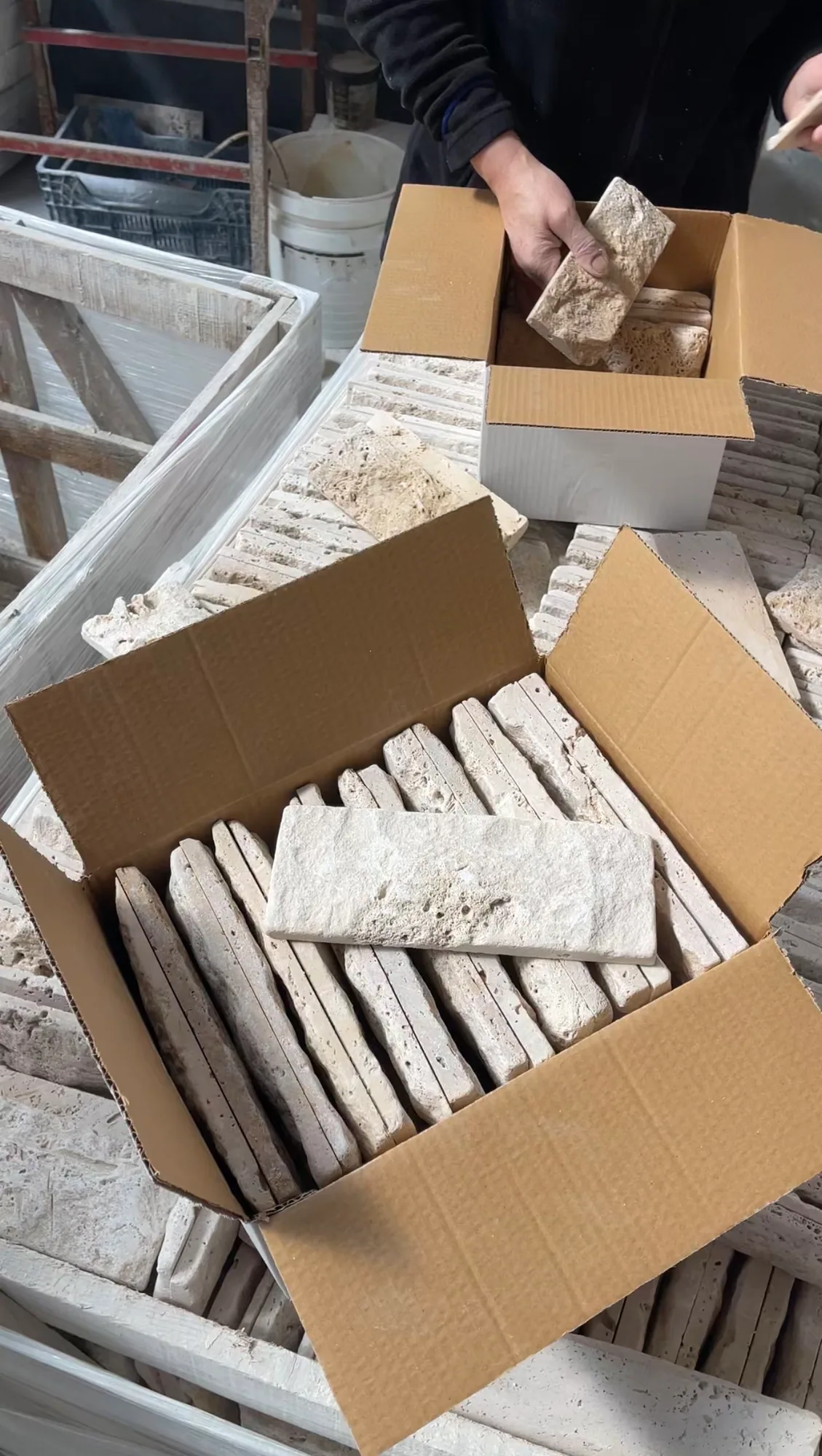 Travertine ledger pieces stacked upright inside open cartons on the packing bench, one piece held above the box as it is loaded.
