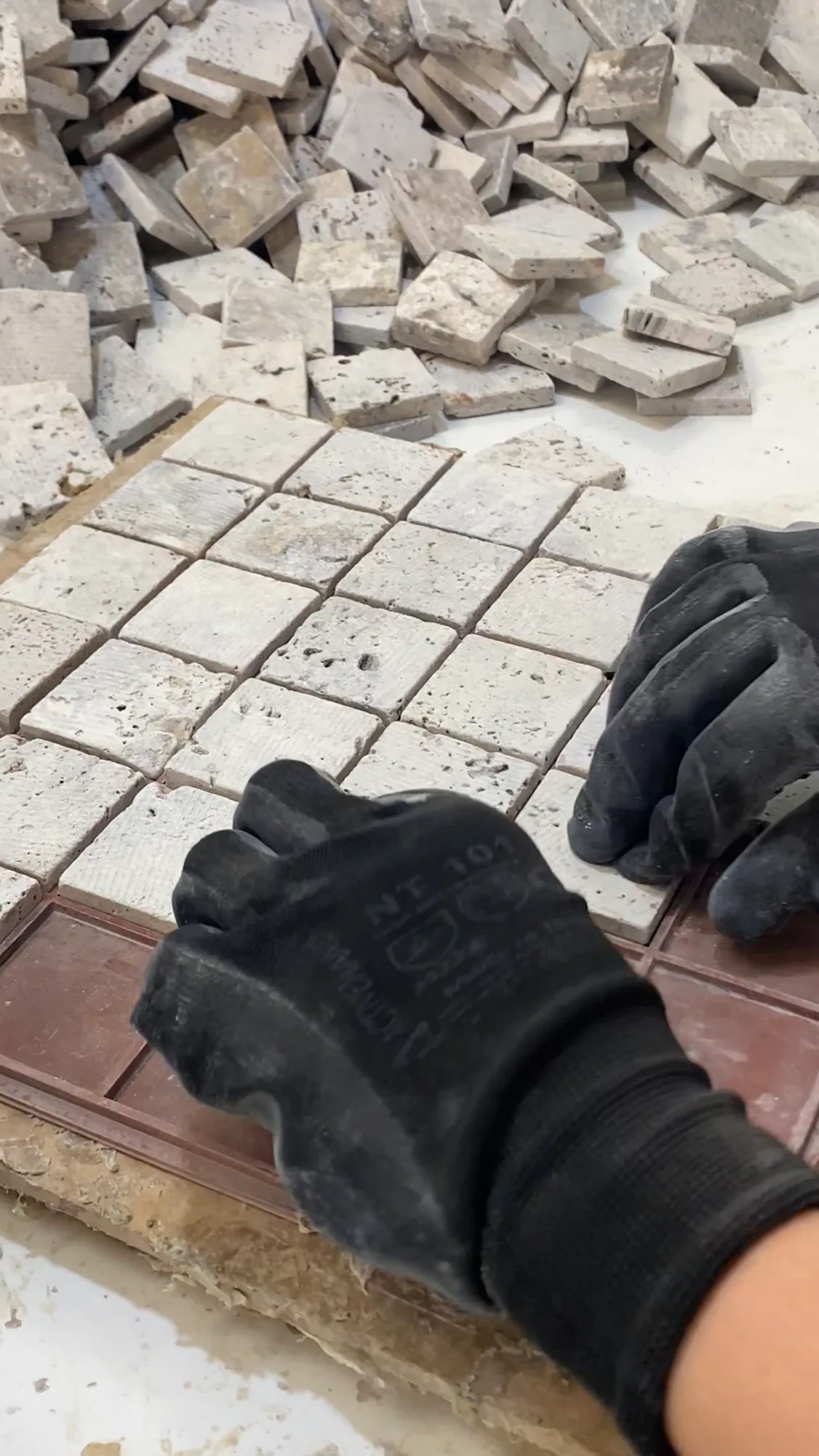 Two gloved hands setting tumbled travertine tesserae into a moulded tray, a loose heap of cut pieces waiting on the bench above.
