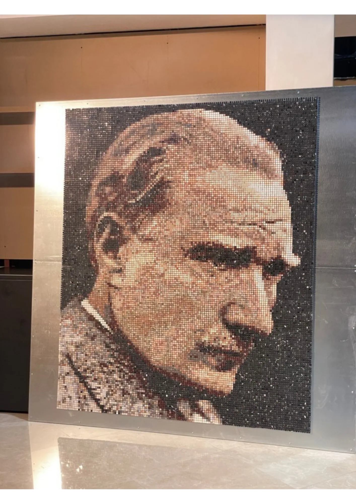 The finished mosaic photographed in place: the portrait panel mounted on a brushed metal backing in a lobby, individual tesserae catching the light unevenly across the face, the polished stone floor below holding a soft reflection.
