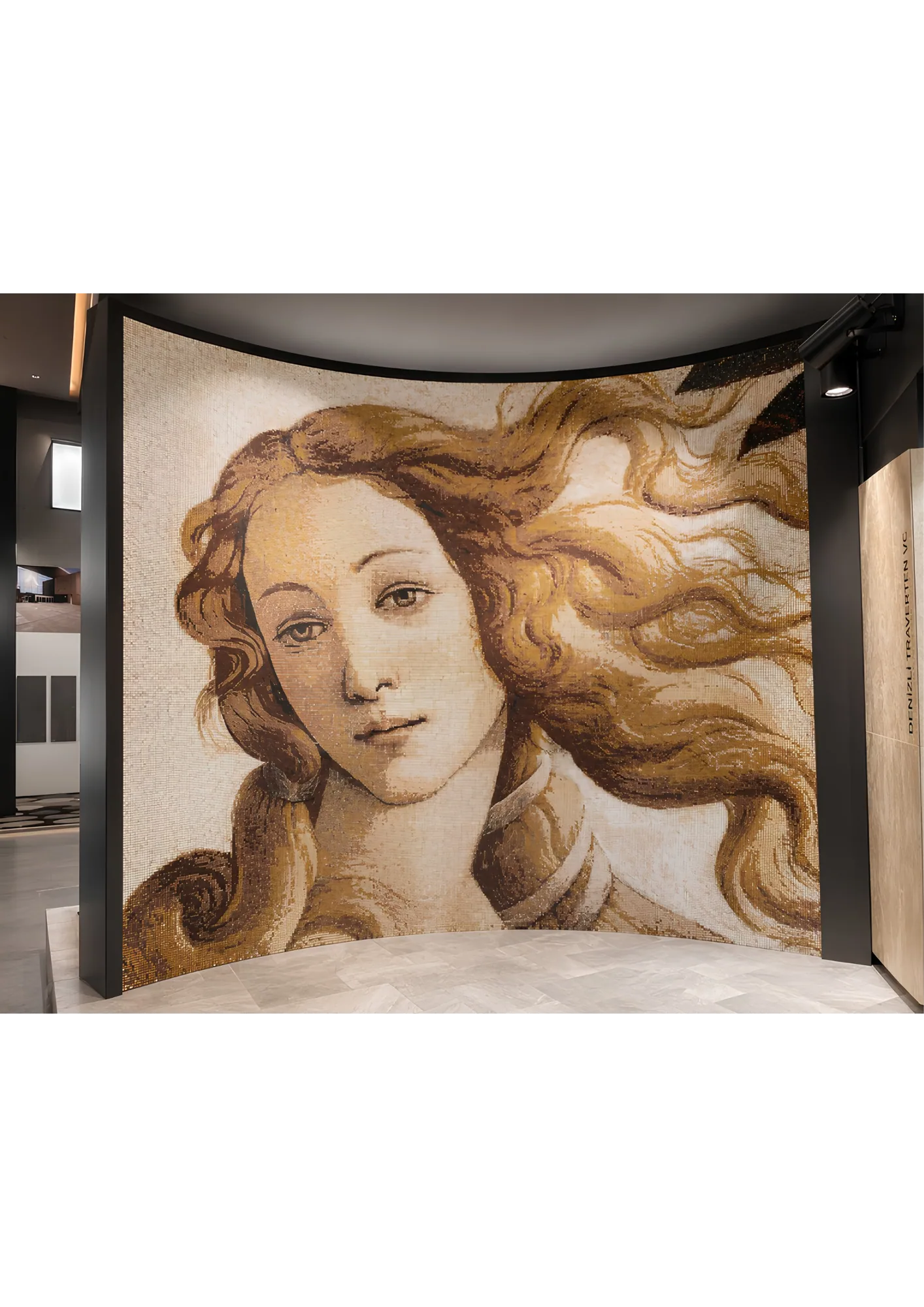 The finished mosaic photographed in place: the head of Venus laid across a tall curved wall in a stone showroom, the tesserae following the flow of her hair and throwing scattered gold glints under two track spotlights, an engraved travertine panel to the right.
